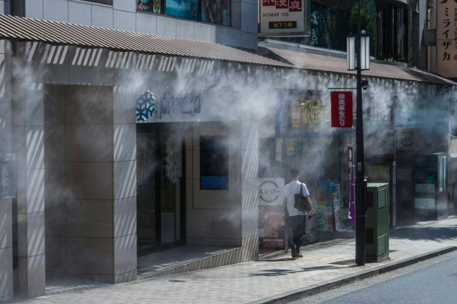 A person walking along a Japanese city sidewalk beside a building with signs and vents releasing a mist of cool vapor.