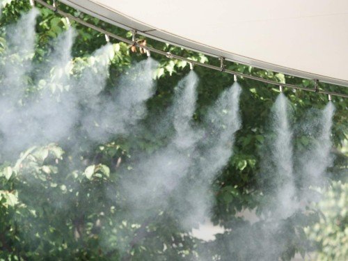 Mist Cooling System installed beneath a canopy, spraying a fine mist to cool an outdoor garden area surrounded by lush green trees.